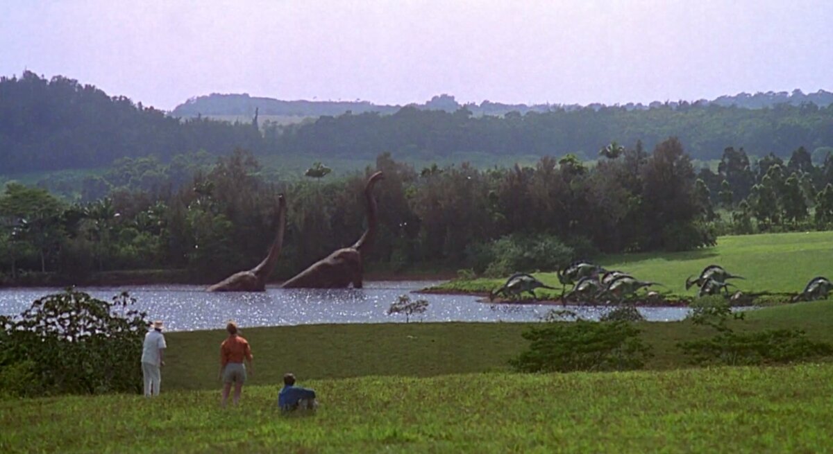 Wide view of dinosaurs roaming the park near a pond